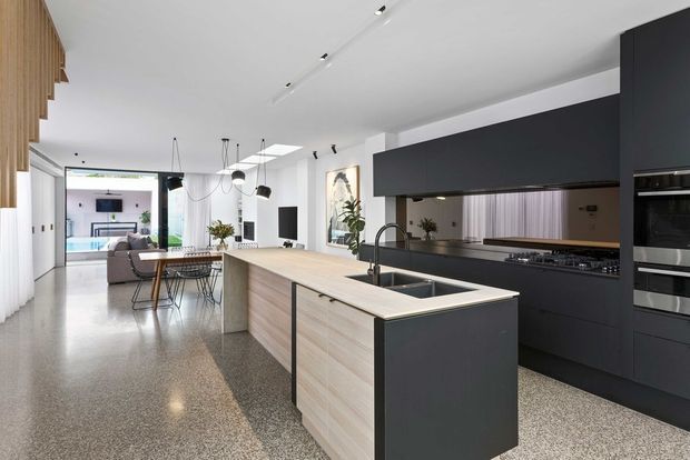 A bright, open-plan contemporary kitchen viewed from the living and dining area. The kitchen features a large island with a pale timber-look finish, dark matte cabinetry, a light benchtop, integrated appliances, a double-bowl sink, and a gas cooktop beneath a dark overhead bulkhead. Polished aggregate-style flooring continues through the space, with white walls, track lighting, pendant lights, and timber screening at the edge of the room. A living area and dining table are visible beyond the kitchen.