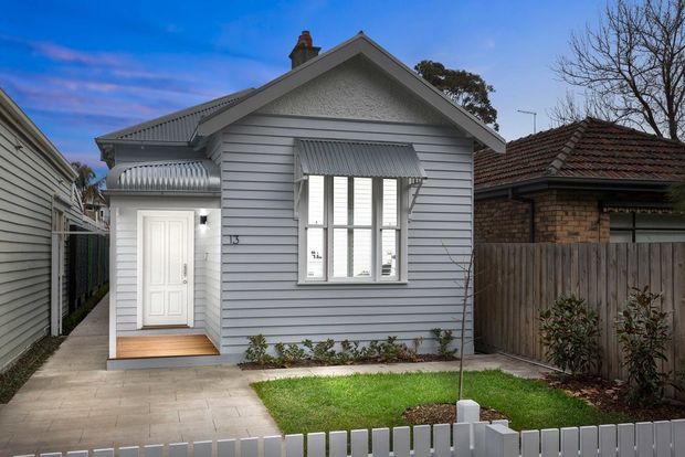 Street-facing view of a compact single-storey cottage with a light grey weatherboard facade, pitched gable roof, white-framed windows, corrugated metal awnings, and a small covered entry. The property has a narrow paved path, compact lawn and low white picket fence, with a neighbouring brick-tiled-roof house visible beside it.