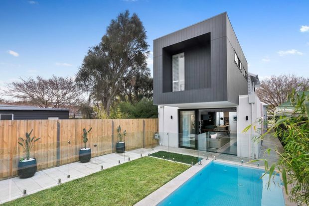 Contemporary two-storey residence with a bold dark vertical-panel upper facade, white lower level, and large glazed opening facing a compact landscaped yard and swimming pool. The property includes a timber boundary fence, paved poolside terrace, lawn, and mature planting under a clear blue sky.