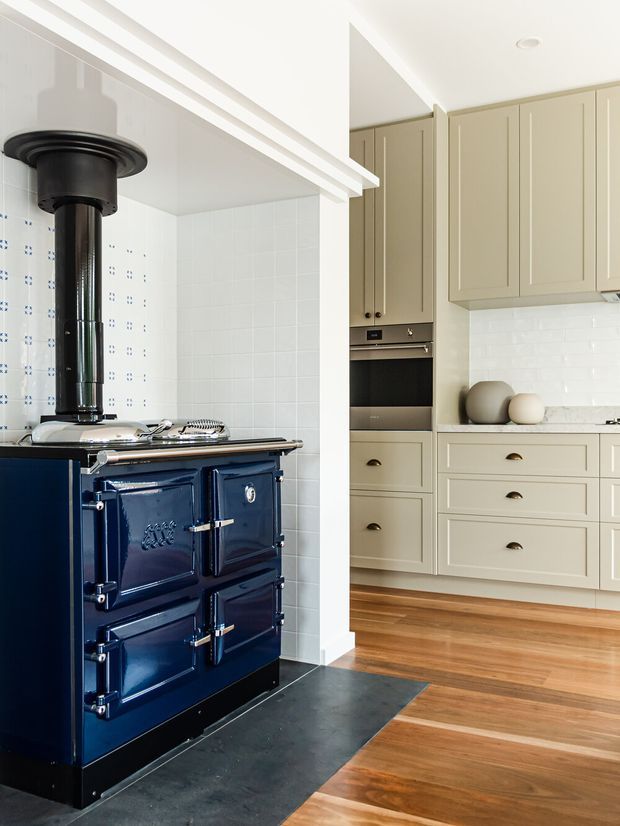 Bright kitchen interior featuring a glossy navy-blue traditional range cooker with a prominent flue, set against white tiled walls. A separate run of pale beige shaker-style cabinetry includes built-in appliances, drawers, brass-toned pulls, and a light benchtop with white subway-tile splashback. Warm timber flooring contrasts with a dark stone or slate hearth in front of the cooker.