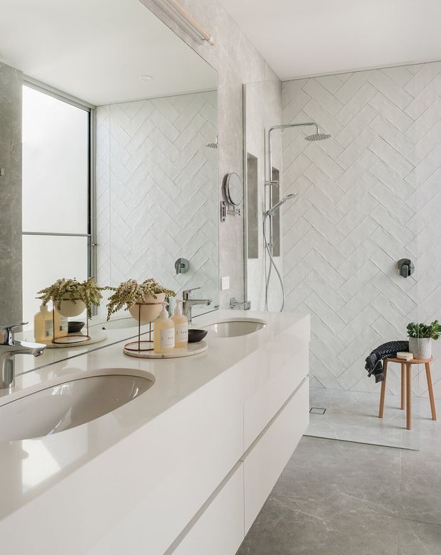 Bright contemporary bathroom with a double floating vanity, integrated white basins, large frameless mirror, chrome tapware and shower fittings, white herringbone wall tiles, and a grey polished concrete-look floor. A walk-in shower occupies the rear of the room, with a frosted window providing natural light and a small timber stool with plant adding warmth.