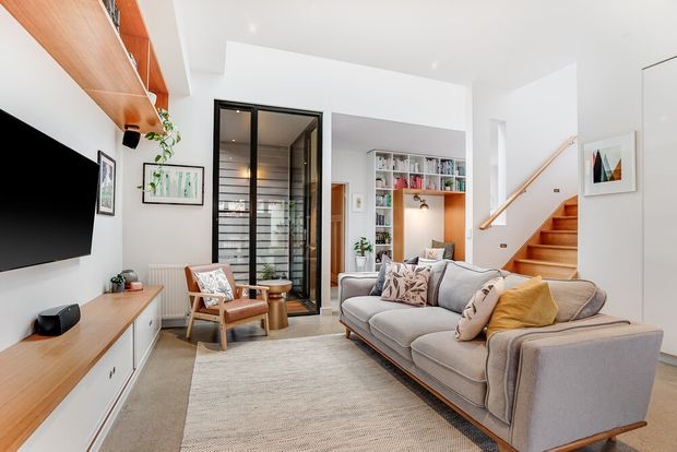 Bright contemporary living room with a large grey upholstered sofa, timber-framed armchairs, a wall-mounted television, built-in timber media cabinetry, shelving, and a compact staircase with timber treads and a timber handrail. Black-framed glazed doors open toward an adjacent space, while a white built-in bookcase and warm timber accents add contrast to the predominantly white interior.