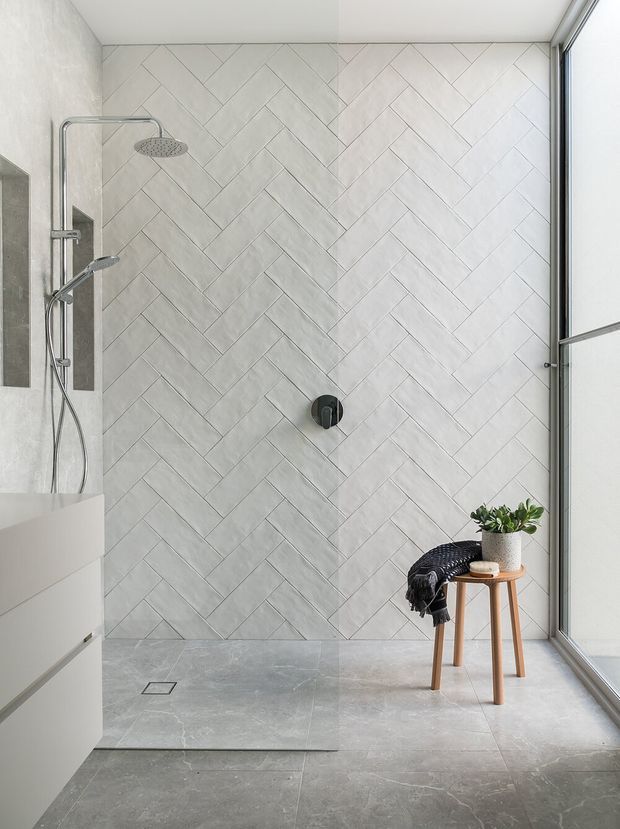 Bright contemporary bathroom featuring a walk-in shower with chrome fixtures, a large light-grey herringbone tiled feature wall, matching grey floor tiles, white built-in cabinetry, and full-height glazing. A small timber stool with a potted plant and folded textile adds warmth.
