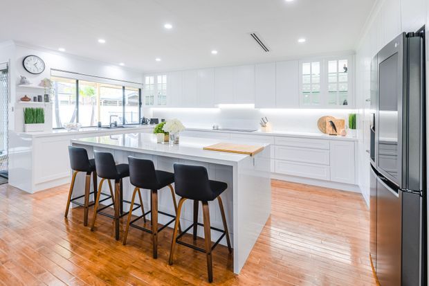 Bright, spacious white kitchen with a large central island, seating for five, extensive white cabinetry, integrated appliances, recessed ceiling lighting, and warm polished timber flooring. Large windows and glass doors bring in abundant natural light.
