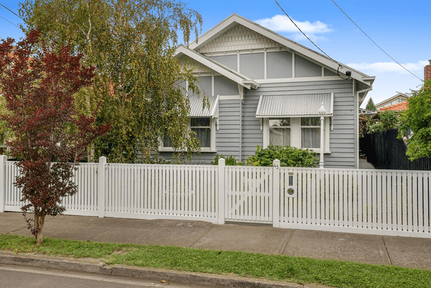 Street-facing view of a traditional single-storey timber house with a prominent front gable, decorative gable detailing, horizontal weatherboard cladding, white trim, and metal awnings. The house is enclosed by a white picket fence and sits in an established suburban streetscape with mature planting.