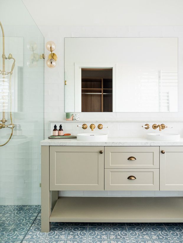 Bright, refined bathroom with a double vanity featuring pale grey-green shaker cabinetry, white countertop, two integrated white basins, and polished brass wall-mounted fixtures. White rectangular wall tiles and a large mirrored cabinet sit above the vanity, with an open timber-lined storage niche visible in the mirror area. A glass-enclosed shower with brass fittings occupies the left side, while patterned blue-and-white encaustic-style floor tiles add a decorative accent. The room has a clean, softly traditional look with warm brass details.