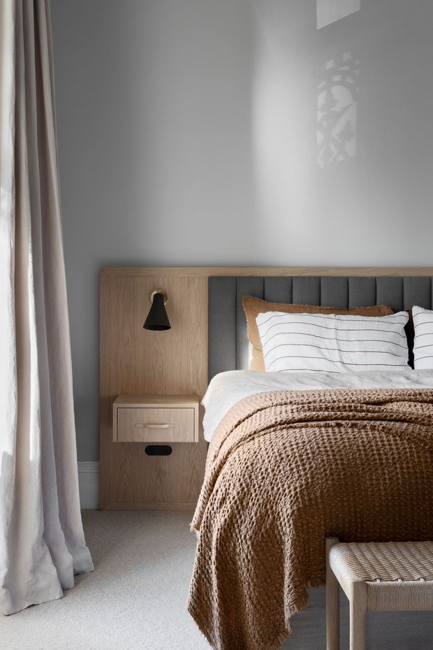 A softly lit contemporary bedroom featuring a wood-panelled integrated bedside unit with a black wall lamp, a dark vertically channelled upholstered headboard, layered white bedding, and a textured warm brown throw. Floor-to-ceiling pale curtains frame the left side, with neutral carpet and a small upholstered bench in the foreground.