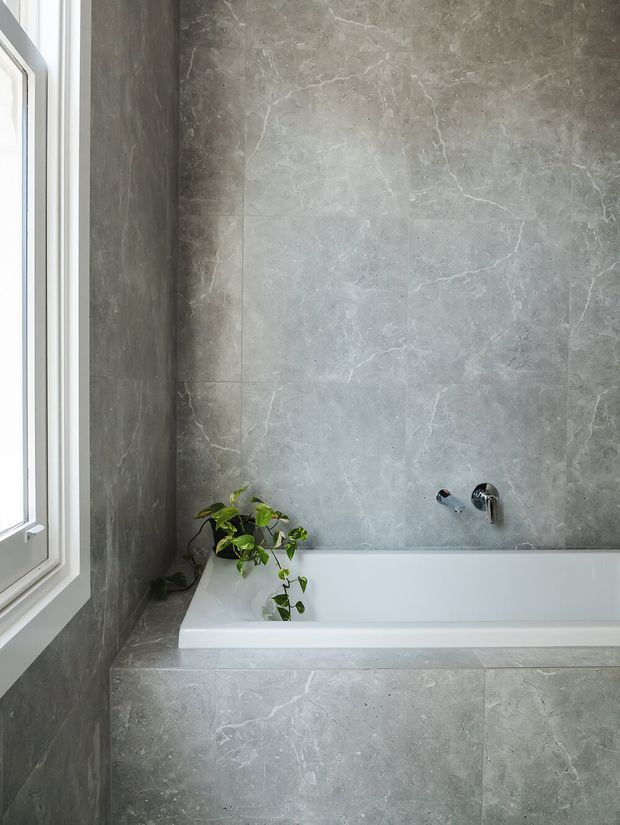 A minimalist bathroom featuring a white built-in bathtub enclosed by large grey marble-effect tiles. A white-framed window brings in natural light, while a small trailing green plant adds a natural accent. Chrome wall-mounted bath fittings are visible above the tub.
