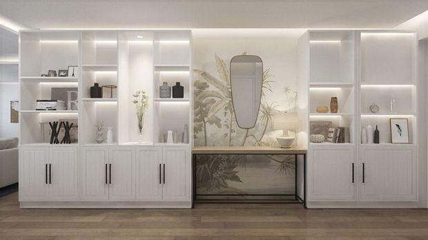 A bright, refined built-in display and storage wall in a residential interior. White cabinetry with recessed-panel doors and black vertical pulls frames illuminated open shelving, while a central dark-framed console with a reflective surface sits beneath a decorative mirror and botanical artwork. The room has a smooth white ceiling, pale walls, and medium-toned timber flooring.