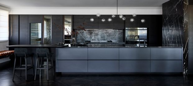 A sophisticated contemporary kitchen with a long dark island and integrated cabinetry, dramatic veined stone backsplash and side wall, dark timber or laminate cabinetry, black flooring, and sculptural pendant lights. Bar stools line the island, while full-height glazing and reflective surfaces add depth.