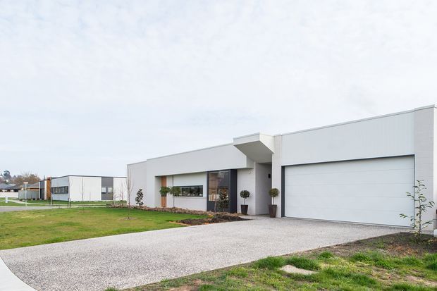Contemporary single-storey residential house viewed from the front, featuring a clean white rendered facade, strong horizontal volumes, a broad integrated double garage, recessed entry, dark-framed glazing, and a pale gravel driveway with a sparsely landscaped lawn.