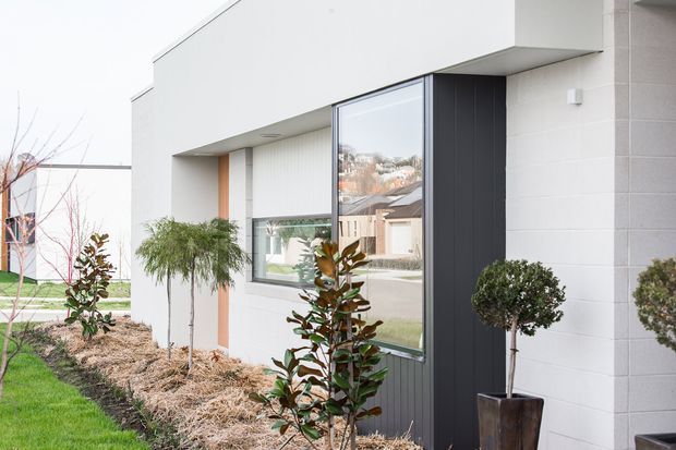 Contemporary minimalist single-storey house facade with crisp white rendered or masonry walls, a projecting flat roof form, large dark-framed windows, and a vertically ribbed dark entry or accent panel. Neatly landscaped planting beds with clipped topiary and young trees soften the geometric exterior. The image is photographed outdoors and focuses on the building envelope, with a residential street and neighbouring houses reflected in the glazing.