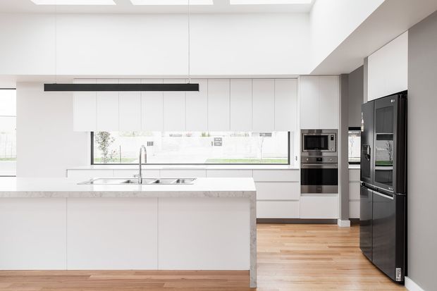 Bright, spacious contemporary kitchen with a large white island, seamless white cabinetry, integrated stainless-steel appliances, black double-door refrigerator, pale stone benchtops, timber flooring, and expansive windows beneath a high ceiling with skylights. A slim linear pendant provides a dark contrast above the island.