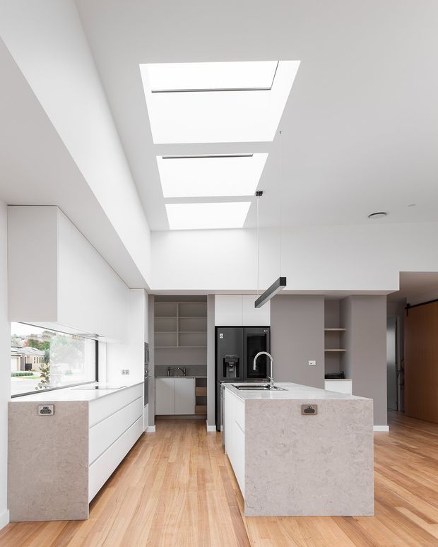Bright contemporary kitchen with a central stone island, integrated sink and curved tap, extensive white cabinetry, open shelving, timber flooring, and a black built-in coffee appliance. Three large skylights and a wide side window provide abundant natural light. The kitchen opens toward a timber-floored circulation or living area.