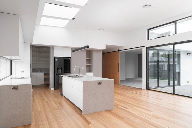 Bright, open-plan contemporary kitchen and living area with a large white island, pale stone benchtops, integrated white cabinetry, timber flooring, black-framed full-height glazing, skylights, and a timber sliding door. The space has a clean, airy, minimalist character and connects directly to an outdoor courtyard through glazed sliding doors.