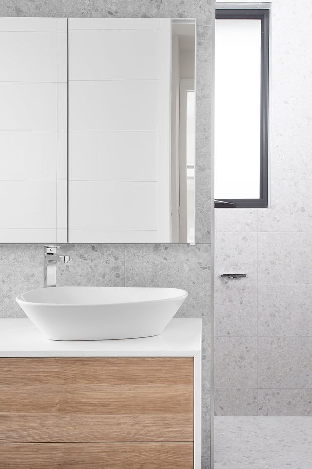 A bright, compact contemporary bathroom featuring a white vessel basin on a timber-look floating vanity, a chrome wall-mounted tap, large-format grey stone-look wall and floor tiles, a mirrored wall cabinet, and a narrow dark-framed window.