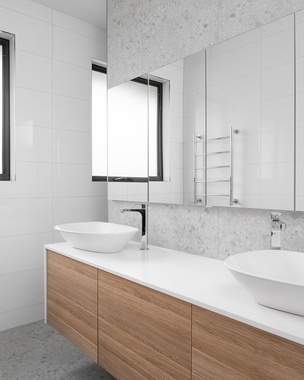 A bright contemporary bathroom featuring a long double vanity with two white vessel basins, a white countertop, and light timber-look drawers. Large-format white wall tiles are paired with grey stone-look feature tiles around the mirror and vanity backsplash. A heated chrome towel rail, wall-mounted chrome taps, frameless mirrored cabinetry, and black-framed windows complete the clean, minimalist space.