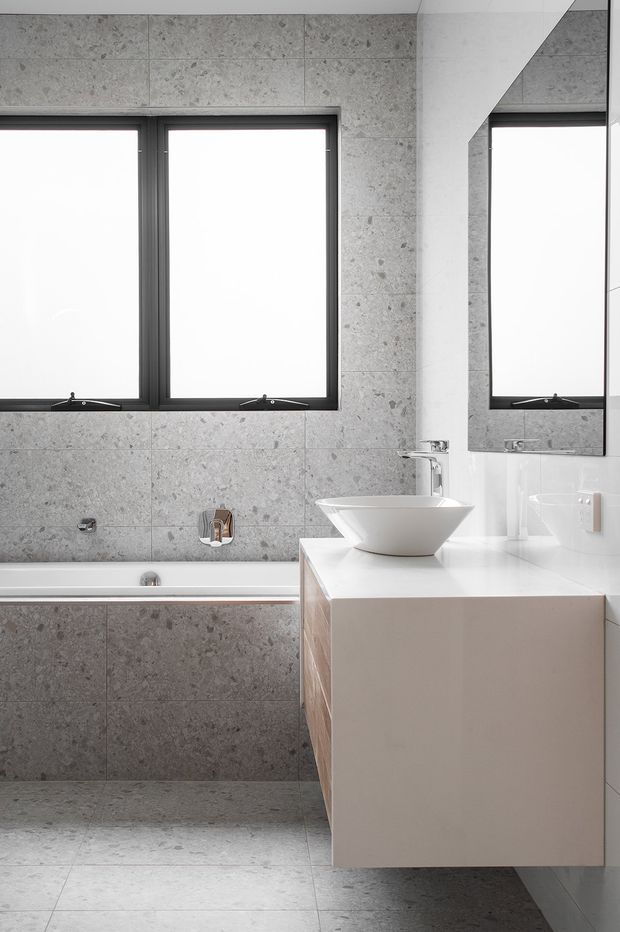 A clean, contemporary bathroom with large grey stone-look tiled walls and floor, a built-in bathtub beneath two dark-framed windows, and a floating pale vanity with a white vessel basin and chrome tapware. A large angular mirror reflects the window and vanity, reinforcing the bright, minimalist composition.