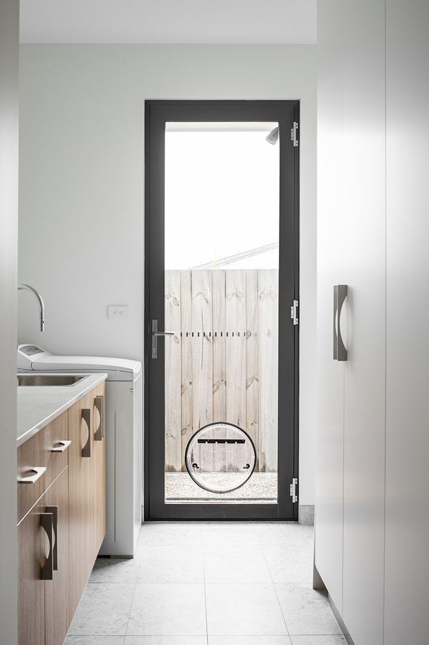 Bright, compact contemporary kitchen or utility space viewed toward a full-height glazed exterior door. The room has pale grey walls, light stone-look tiled flooring, light timber-grain cabinetry with dark metal handles, a pale benchtop, stainless tapware, and integrated appliances. The black-framed door opens to a small fenced outdoor area, while a large curved wall handle or accessory is mounted beside it.