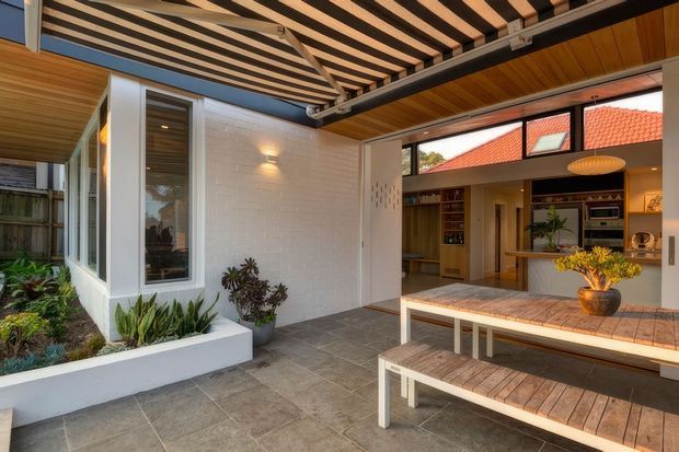 A covered indoor-outdoor entertaining area with a timber slatted pergola and timber-lined ceiling, opening into a contemporary kitchen and living space. The courtyard features large grey floor tiles, a built-in white planter with greenery, and a substantial timber dining table with matching bench seating. The interior beyond includes timber cabinetry, open shelving, a kitchen island, and clerestory-style glazing. Warm natural timber contrasts with white masonry and dark metal framing.