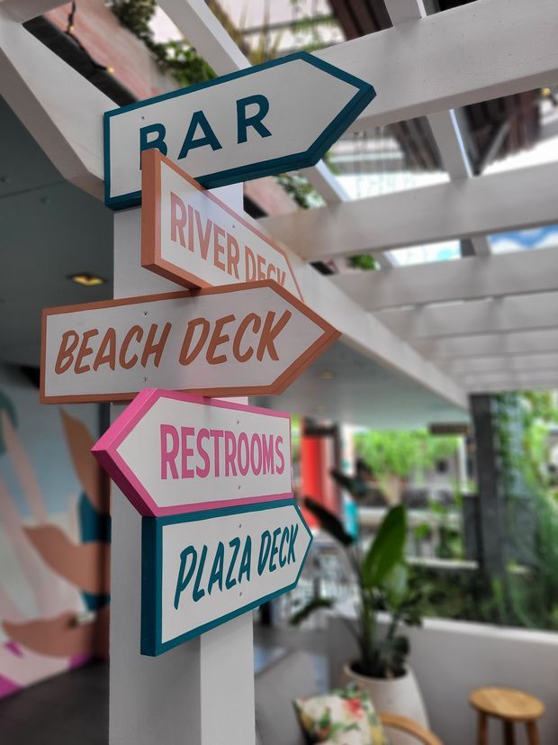 A colorful directional signpost inside a bright, open-air commercial venue. Arrow signs point toward a bar, river deck, beach deck, restrooms, and plaza deck. The space features a white exposed-beam canopy, indoor greenery, and casual seating in the background.