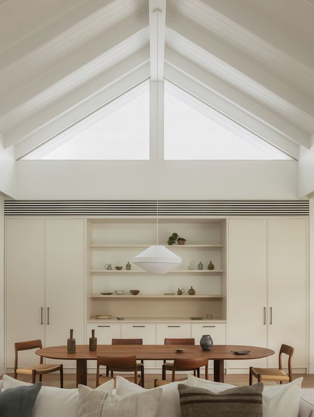 A bright, refined open-plan dining area with a long oval timber dining table and matching wooden chairs, viewed from behind a sofa. The room features extensive off-white built-in cabinetry and open display shelving, a minimalist white pendant light, and a dramatic pitched ceiling with exposed white-painted beams and high triangular clerestory glazing. The styling is warm and contemporary with understated ceramic and decorative objects.