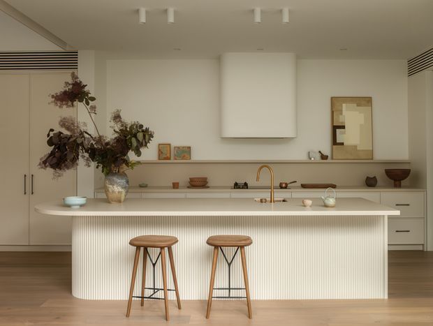 A warm, refined contemporary kitchen with a large sculptural island, vertically fluted island frontage, integrated pale cabinetry, timber flooring, and a simple white rangehood. Two timber bar stools sit at the island, while restrained artwork, ceramics, and dried botanical arrangements add organic detail. The space is softly lit with a neutral beige and cream palette and brass-toned tapware.
