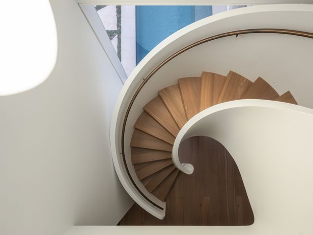 A sculptural spiral staircase viewed from above, with warm timber treads radiating around a central curved void. The staircase has smooth white curved walls and balustrades, a dark bronze-toned handrail, and a narrow skylight or glazed opening above that brings in daylight. The composition is highly geometric and contemporary, with a refined, minimalist architectural character.