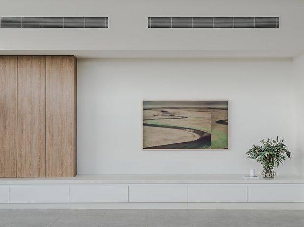 Minimal, light-toned interior wall with extensive built-in cabinetry and a long low white joinery unit. Full-height light timber cabinet doors occupy the left side, while the white wall features a framed landscape artwork and a small potted plant. Linear ceiling air-conditioning grilles and pale large-format flooring reinforce the clean contemporary design.