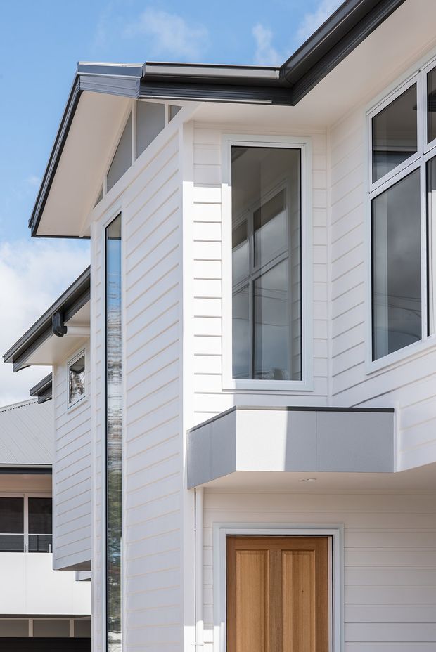 Close-up exterior view of a contemporary multi-storey residential facade. The building features white horizontal weatherboard cladding, dark grey roof edge and gutters, white-framed windows, a tall narrow glazed opening, and a projecting grey-clad balcony or entry canopy above a timber front door. Bright daylight emphasizes the clean, crisp facade composition.