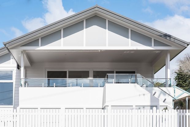 Front exterior view of a bright white, two-storey residential house with a prominent gabled roof, deep covered upper balcony, glass balustrades, white vertical fencing, and clean contemporary detailing. The facade combines white cladding with pale grey gable infill and dark-framed glazing.