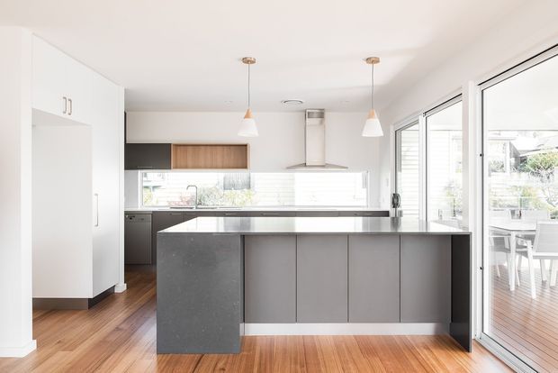 Bright contemporary kitchen viewed from the living area, featuring a large central island with a dark stone benchtop and grey flat-panel cabinetry, white full-height pantry cabinetry, timber flooring, and a compact linear kitchen with integrated appliances. White pendant lights hang above the island, while extensive windows and glazed doors provide natural light and connect to an outdoor dining deck.