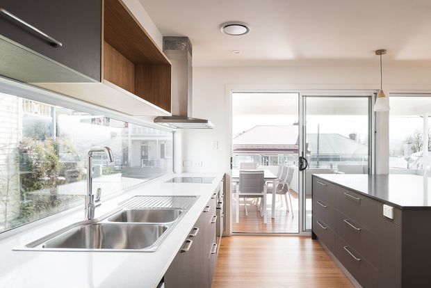 Bright contemporary kitchen with a long white benchtop, double stainless-steel sink, dark lower cabinetry, timber open shelving, and a central dark island. Full-height glazing and sliding glass doors open to a dining area and provide abundant natural light. Timber flooring continues through the kitchen and adjacent dining space.