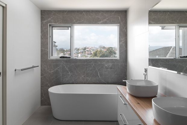 Modern bathroom with a freestanding white bathtub, twin vessel basins on a timber-look vanity, large wall mirror, and a grey marble-effect tiled feature wall. A wide window provides natural light and views over nearby homes.