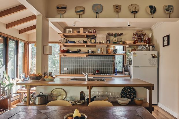 Warm, characterful open-plan kitchen viewed from the dining area. The room features a large timber island with open shelving, a muted green small-format tiled splashback, timber flooring, exposed timber ceiling beams, open timber shelving filled with cookware and decorative objects, and a white refrigerator. Tall glazing at the left brings in abundant natural light and overlooks greenery. A dining table occupies the foreground.