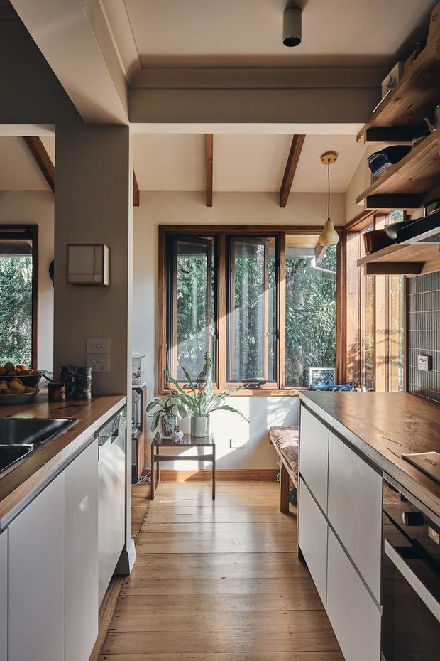 A compact, galley-style kitchen viewed down the central walkway toward a bright window nook. The space features warm timber flooring, exposed timber ceiling beams, wood-framed glazing, white flat-panel cabinetry, dark benchtops, and open timber shelving. A small built-in bench and potted plant sit by the windows, creating a warm, naturally lit interior.