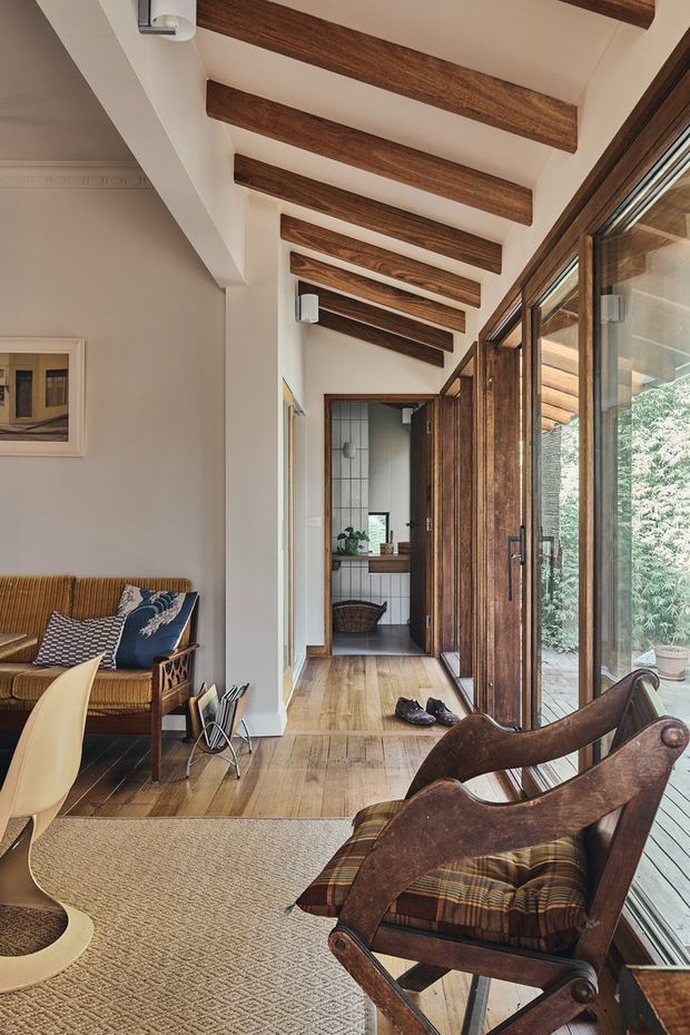 A warm, narrow interior corridor and living area with full-height timber-framed glazing along one side, opening toward an outdoor deck. The space features exposed dark timber ceiling beams, white plaster walls, wide timber flooring, a woven neutral rug, and vintage wooden lounge chairs with patterned upholstery. A compact tiled bathroom or utility space is visible at the end of the passage. The room has abundant natural light and a relaxed, rustic-modern character.