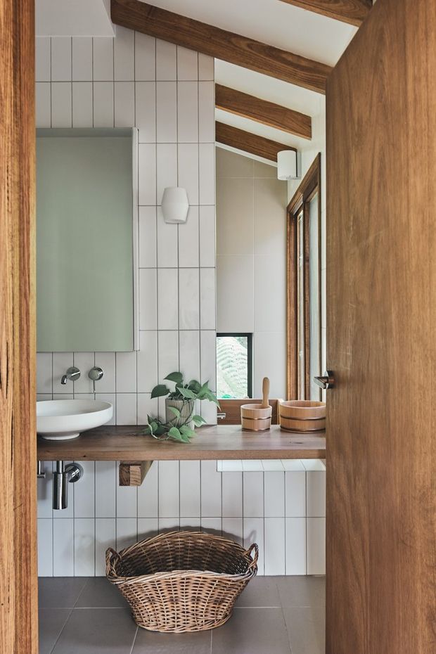 A compact, warmly styled bathroom viewed through an open timber door. The room features a wall-mounted white vessel basin on a dark timber vanity shelf, a large frameless mirror, vertically arranged white tiles with dark grout, a frosted glass shower screen, timber-framed windows, exposed dark timber ceiling beams, and a woven basket beneath the vanity. Natural wood, greenery, and simple ceramic accessories create an inviting contemporary-rustic feel.