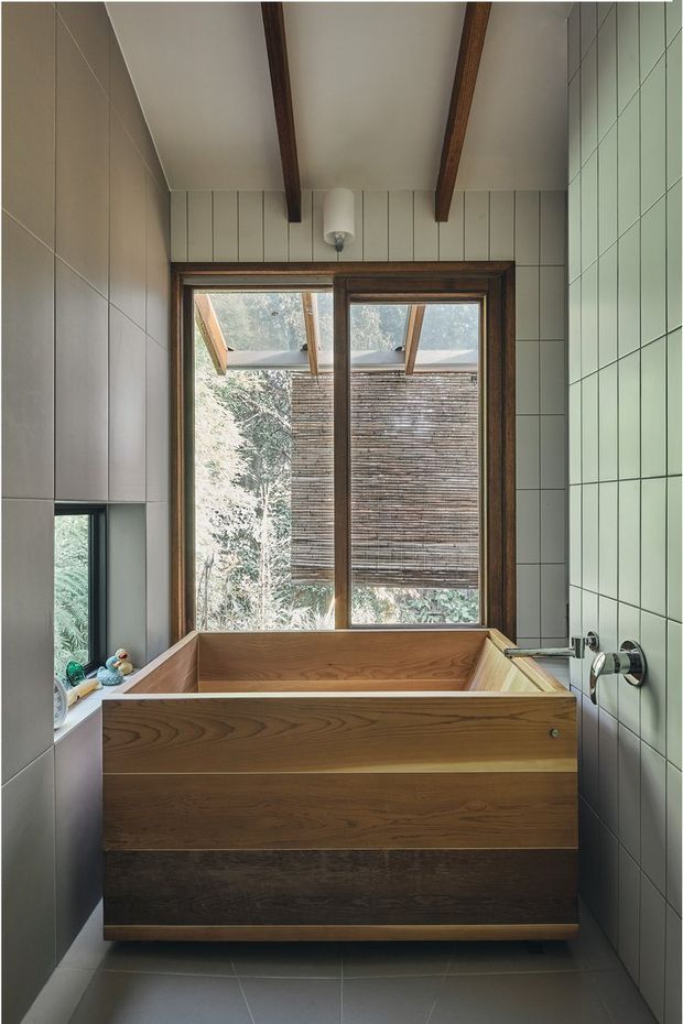 A compact, refined bathroom centred on a freestanding timber-clad bathtub beneath a large timber-framed window. Pale square wall tiles, dark wood ceiling beams, a wall-mounted chrome tap, and a narrow recessed shelf contribute to the warm, natural aesthetic. The window overlooks leafy vegetation and a screened exterior structure.