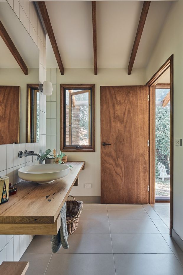 A warm, contemporary bathroom with a floating timber vanity, round vessel basin, wall-mounted black tapware, and large pale square floor and wall tiles. A full-height mirror reflects the vanity area, while exposed dark timber ceiling beams, timber-framed windows, and a timber door add architectural character. Natural light enters through the window and open door, with greenery visible outside.