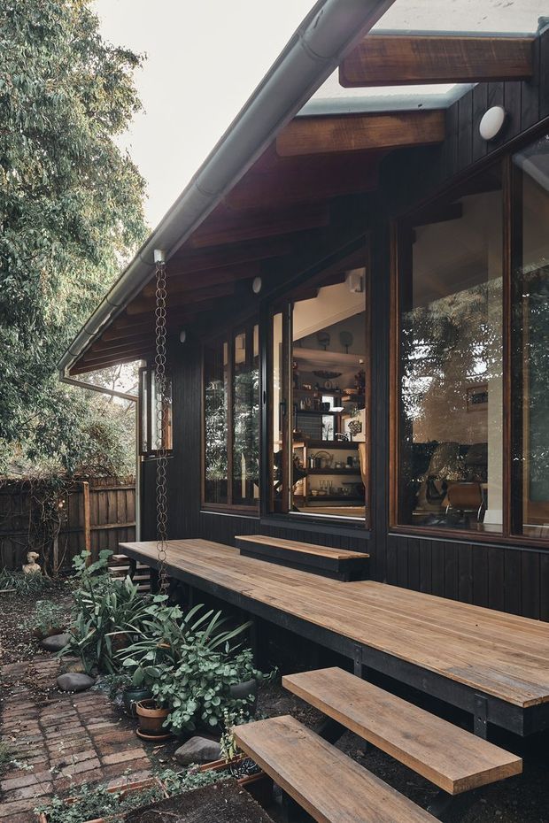 Exterior view of a compact timber-clad home with a deep eave, exposed dark timber beams, large framed windows, and a long timber deck with integrated bench seating and steps. A hanging rain chain, brick paving, planted garden beds, and mature trees surround the outdoor entertaining area. The interior is visible through the glazing but the exterior deck and facade dominate the image.