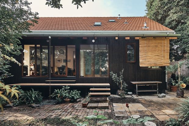 Single-storey dark-clad house viewed from a landscaped paved garden courtyard. The facade has vertical dark timber cladding, broad timber-framed glazing, a low-pitched terracotta tiled roof with skylights, a covered entry porch, exterior steps, and a rolled bamboo blind over one window. Brick and concrete paving, planted garden beds, and a simple outdoor bench create a warm, natural residential setting.