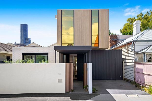 Street-facing view of a narrow contemporary infill residence, with a vertically proportioned upper storey clad in warm timber-toned vertical boards, tall dark-framed windows, and a dark recessed entry and garage frontage. A low rendered boundary wall and pedestrian gate define the front setback, while older neighbouring homes flank the modern addition. A high-rise tower is visible in the distant urban background.