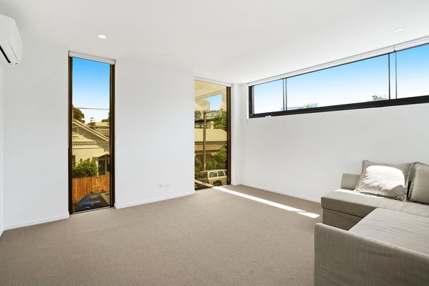 Bright contemporary living room with white walls and ceiling, beige carpet, expansive horizontal clerestory-style windows, a narrow full-height window, and a light grey sectional sofa. Strong natural light enters from the glazing, with neighbouring residential rooftops visible outside.