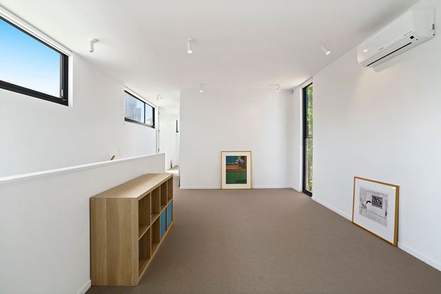 Bright, minimalist interior room with white walls and ceiling, beige carpet, narrow black-framed windows, a low timber built-in shelving unit, framed artwork, and a wall-mounted split-system air conditioner. The long rectangular space is sparsely furnished and appears to function as a living or multipurpose room.
