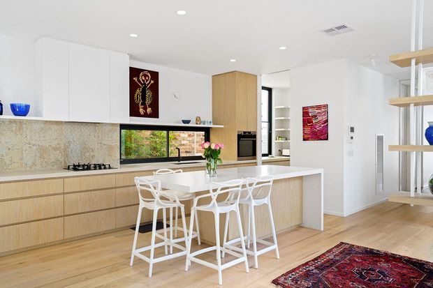 Bright contemporary open-plan kitchen with a large central island and four white bar stools. The space features pale timber cabinetry and flooring, white overhead cabinetry and walls, a textured stone-look splashback, integrated appliances, black-framed horizontal window glazing, and open timber shelving at the right. Colourful artwork, blue glassware, flowers, and a patterned rug add accents.