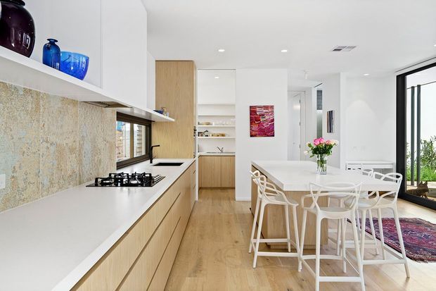 Bright open-plan kitchen and dining area with light timber cabinetry, white walls and benchtops, a textured stone-look splashback, and a white dining table with modern white chairs. Large glazing brings in natural light and greenery is visible outside.