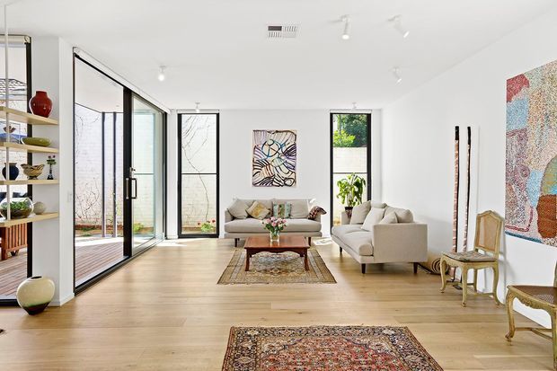 Bright, spacious contemporary living room with white walls, pale timber flooring, large black-framed glazed doors and windows, open shelving, neutral sofas, vintage occasional chairs, patterned rugs, and colourful wall art. The room opens toward a small courtyard or garden area through full-height glazing.