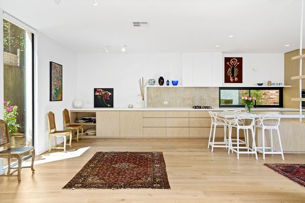 Bright, open-plan contemporary kitchen and dining area with white walls and cabinetry, pale timber flooring, a long light-wood kitchen island with integrated seating, and a textured neutral stone or concrete-look splashback. Large glazing brings in garden views, while Persian-style rugs, artwork, plants, and sculptural objects add warmth and colour. A compact study nook is integrated into the kitchen cabinetry.
