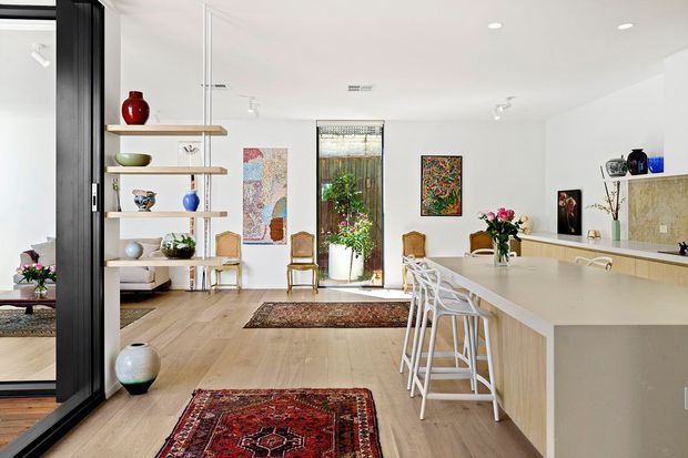 Bright open-plan contemporary kitchen and living area with pale timber flooring, a large light-coloured island, white bar stools, open display shelving, framed artwork, indoor plants, and full-height black-framed glazing. The space has abundant natural light and a clean, refined yet warm atmosphere.